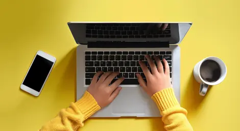 Birdseye view of someone using a laptop with a coffee and mobile phone next to them on a yellow table